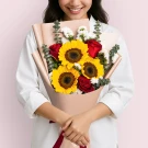Woman holding a bouquet of sunflowers, red roses, and white statice, wrapped in pink paper.