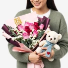 Pink and red roses with eucalyptus, a "Best Mom Ever" cake topper, and a teddy bear wearing a blue scarf with a red heart.