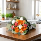 A bright bouquet of orange gerberas, carnations, and white flowers wrapped in green paper with a white ribbon.