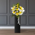 Yellow gerberas and purple pingpong mums in a white bouquet on a black stand with white ribbon.