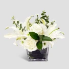 White roses, lilies, and daisies in a clear square vase with greenery.