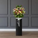 Flower arrangement with white lilies, pink roses, yellow and white carnations, yellow mums, and white matthiola on a black stand with a silver bow.