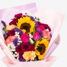 Vibrant bouquet with sunflowers, pink roses, carnations, mums, and statice, wrapped in pale pink paper.