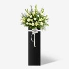 White lilies, carnations, roses, and matthiola in a tall black stand with a silver bow.