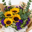 A bouquet of sunflowers, purple statice, eucalyptus, and white filler flowers wrapped in brown paper and white fabric.