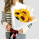 Woman holding a bouquet of 4 sunflowers wrapped in white paper, tied with a red and black ribbon.
