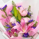 Pink stargazer lilies, pink roses, and purple statice flowers in a bouquet with pink and white paper wrapping.