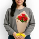 Woman holding a bouquet of three red roses wrapped in brown paper, with a yellow ribbon.