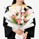 A person in graduation attire holds a bouquet of white anthuriums, pink tulips, white roses, and pink carnations.
