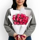 A person holds a bouquet of 18 pink roses and white accents, wrapped in white paper and tied with a burgundy ribbon.