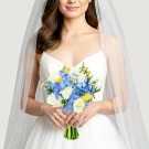 Bride in a white dress holding a bouquet of white roses, blue hydrangeas, and yellow tulips under a veil.
