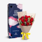 A bouquet of 12 red roses with eucalyptus, wrapped in brown paper and tied with a yellow ribbon, next to a blue gift box.