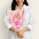 A woman in a white shirt holds a pink bouquet of dried flowers, including a rose, on a pale background.