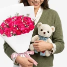 Woman in green sweater holding pink roses bouquet and white teddy bear with blue scarf.