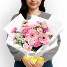 Woman holding a pink gerbera and carnation bouquet wrapped in white paper, tied with a yellow ribbon.