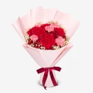A bouquet of red and pink carnations with baby's breath, wrapped in pale pink paper and tied with a dark red ribbon.
