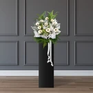 White gerbera daisies and greenery on a black pedestal with white ribbon, against a dark grey paneled wall.