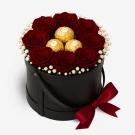 Round black box with preserved red roses, golden chocolates, and a burgundy bow.