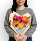 Woman holding a bouquet of orange gerbera daisies and pink roses, wrapped in peach and gray paper with a pink ribbon.