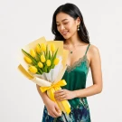 A person holds a bouquet of 10 bright yellow tulips with green leaves, wrapped in cream and yellow paper, tied with a yellow ribbon.