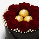A round black box filled with preserved red roses and three Ferrero Rocher chocolates.