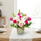A square glass vase holds a vibrant floral arrangement of pink roses, white lilies, and pink carnations.