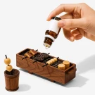 Miniature wooden grill with skewers of food and a hand dripping oil from a bottle.