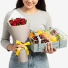 Woman holding a bouquet of red roses and a clear fruit basket with oranges, lemons, grapes, and apples.
