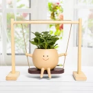 A happy face plant pot with arms and legs sits on a swing, holding a green plant.