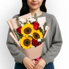 Woman holding a bouquet of sunflowers, red roses, and white statice flowers, wrapped in blush paper.