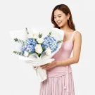 A woman holds a bouquet of blue hydrangeas, white roses, and white carnations, elegantly wrapped in white paper.