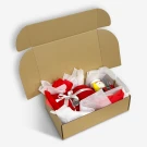 Open cardboard gift box filled with red and white tissue paper, a red ornament, and a Santa Claus figurine.
