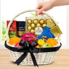 A white wicker basket filled with Ferrero Rocher chocolates, oranges, a rose, and other assorted treats, with a hand holding the handle.