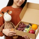 Person holding a gift box containing a plush giraffe, grapes, apple, orange, and a bottle of water.