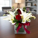 A lush arrangement of white lilies, red roses, and white chrysanthemums in a glass vase with a red ribbon.