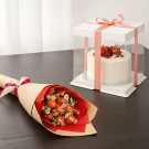 White cake with berries in a clear box next to a bouquet of strawberries and daisies wrapped in brown paper.