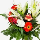 A bright floral arrangement of white lilies, red gerbera daisies, red carnations, and white roses with green ferns.