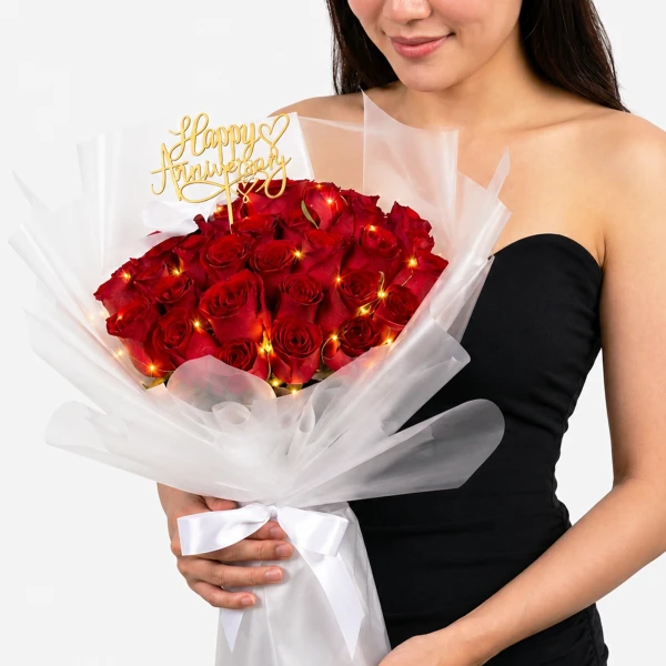 A woman in a black dress holds a large bouquet of red roses, adorned with fairy lights and a gold "Happy Anniversary" topper.