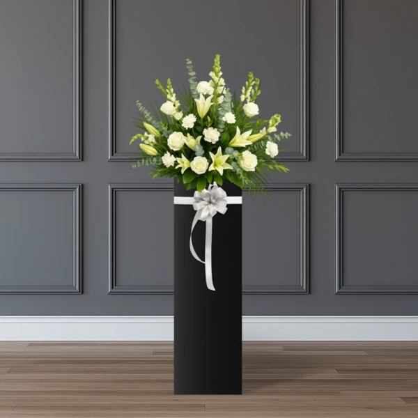 A tall black stand holds a white flower arrangement with lilies, roses, and carnations, adorned with a silver bow.