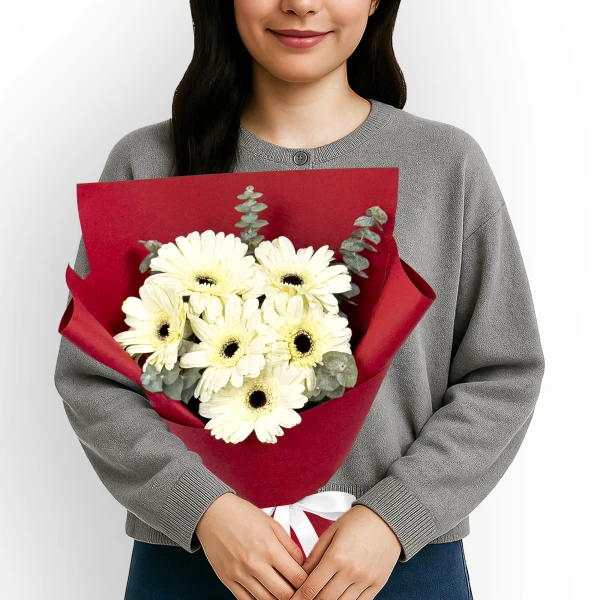 White gerberas and eucalyptus bouquet wrapped in red paper, held by a person in a gray sweater.
