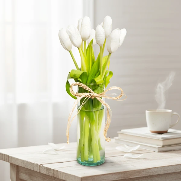 White tulips in a glass vase with a twine bow on a wooden table, with a steaming mug and books in the background.