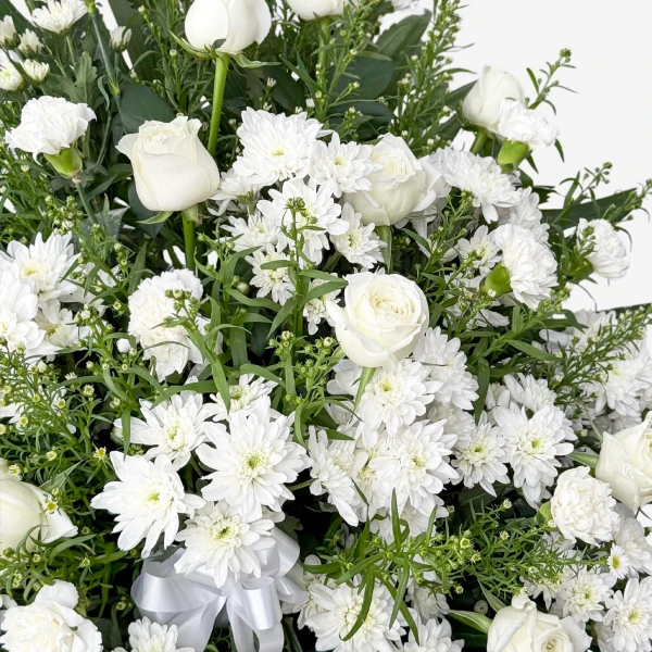 White roses, mums, and carnations arranged in a sympathy stand with greenery and a white ribbon.