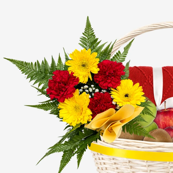 Basket of red and yellow gerbera daisies, red carnations, ferns, fruit, and a red knitted item.