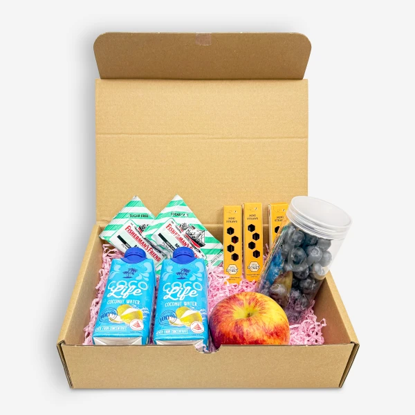 Cardboard box filled with wellness treats: coconut water, Fisherman's Friend, honey straws, blueberries, and an apple.
