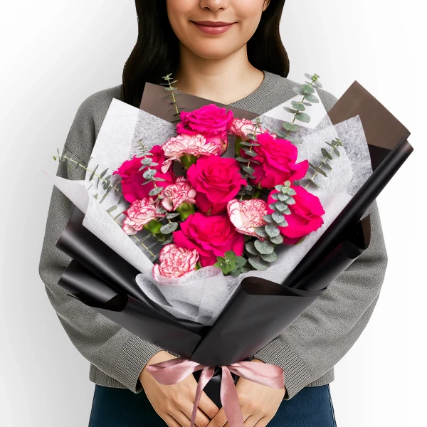 Woman holding a bouquet of bright pink roses, pink carnations, and eucalyptus leaves, wrapped in black and white paper with a pink ribbon.
