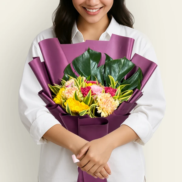 Woman in white shirt holding a bouquet of yellow, pink, and fuchsia carnations and roses, wrapped in purple paper.
