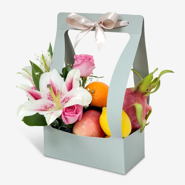 Gift basket with pink roses, lilies, orange, apple, lemon, and dragon fruit, tied with a bow.