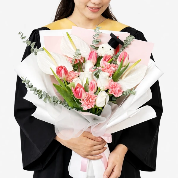 A person in graduation attire holds a bouquet of white anthuriums, pink tulips, white roses, and pink carnations.