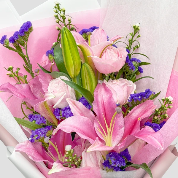 Pink stargazer lilies, pink roses, and purple statice flowers in a bouquet with pink and white paper wrapping.