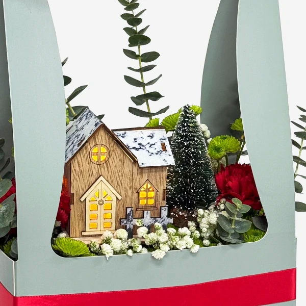 A miniature wooden house, Christmas tree, and festive flowers in a decorative box.